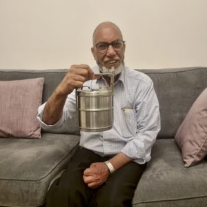 A coloured photograph of a British Asian man sat on a sofa, smiling. He is holding the tiffin box he was gifted by his Father.