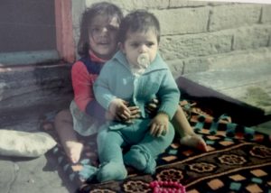 A coloured photograph of a child sat with a baby on her lap outside their house, on a rug. Photograph: Mohammed Ashraf