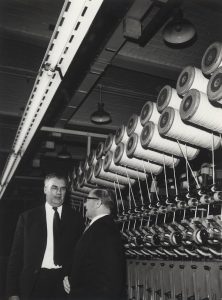 A black and white photograph of the drawing machinery and two men in suits at Salts Mill. Photograph: Bradford Museums & Galleries