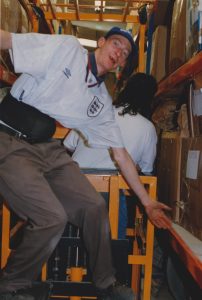 A portrait, coloured photograph of a packer in a football shirt standing on machinery in a warehouse. Photograph: Richard Smith