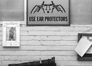 A black and white photograph of a sign saying 'Use Ear Protectors' with an illustration of someone with their fingers in their ears, plus a calendar and noticeboard, in the weaving shed of Drummonds Mill, 1991. 